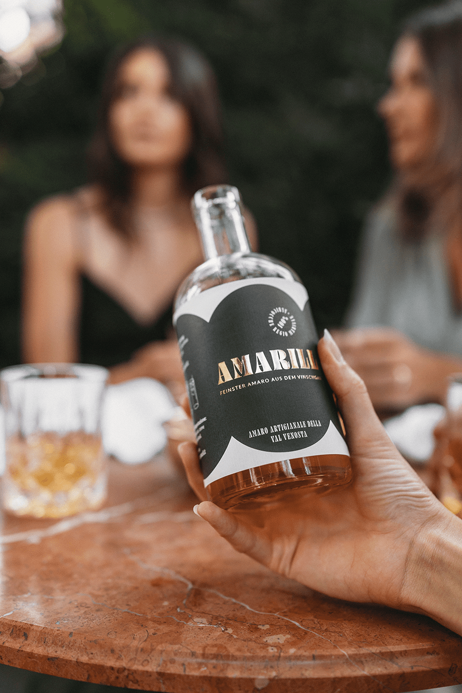 Eine Person hält eine Flasche Amaro Amarill in der Hand, im Hintergrund sitzen mehrere Menschen mit einem Glas Amaro Amarill am Tisch.